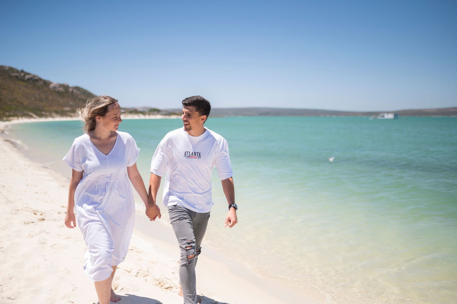 Couple holding hands walking on a sunny white sand beach with turquoise water in the background.