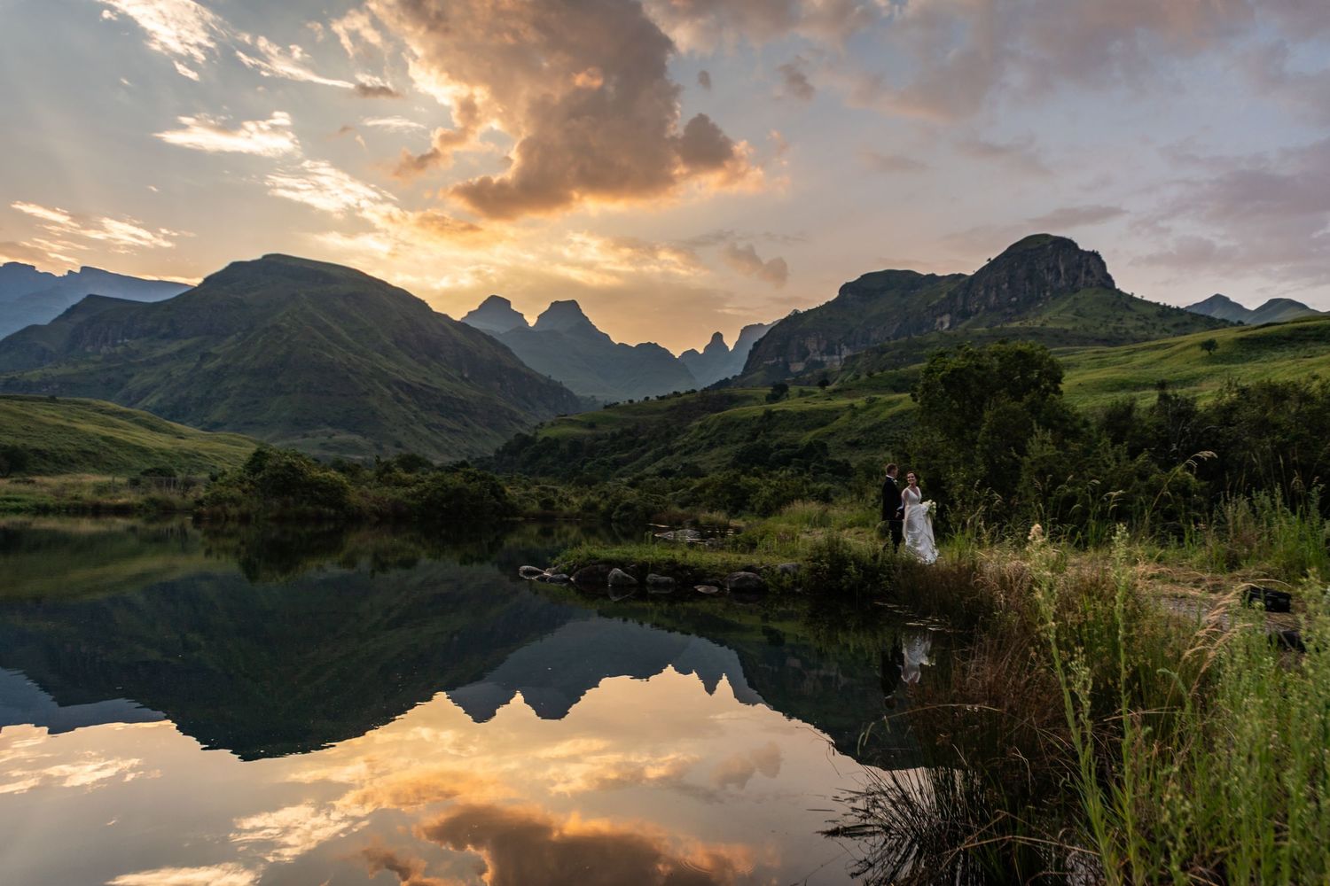 Couple stands by a reflective lake at sunset with dramatic mountain peaks and glowing clouds in a lush green valley.