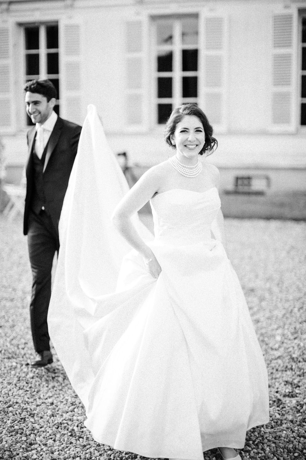 Bride smiling with groom walking behind her at Château Barthélemy wedding near Paris