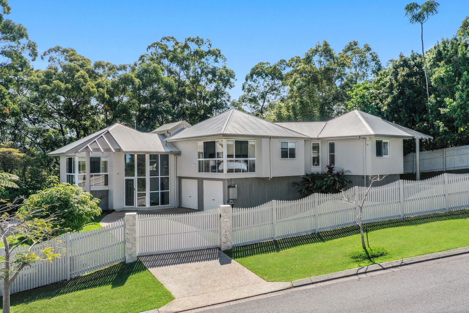 White rendered family home with gabled roof, white picket fence, and tropical garden setting in bright daylight.