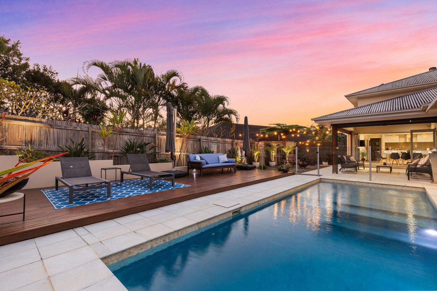 Luxurious backyard pool with timber deck, outdoor lounge seating, palm trees, and vibrant sunset backdrop.