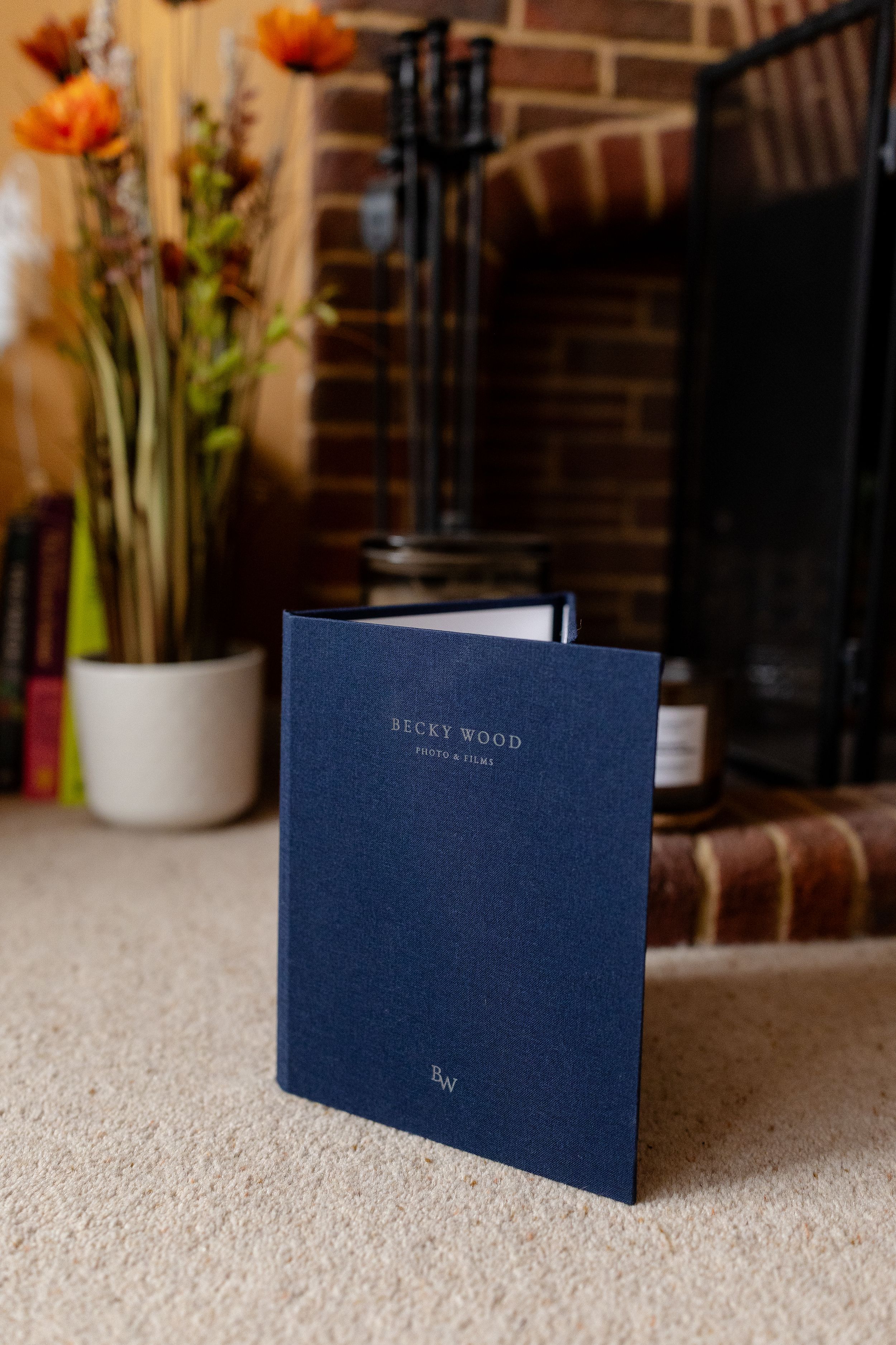 Navy blue wedding album book shown from different angles on white carpet with floral decor in background.