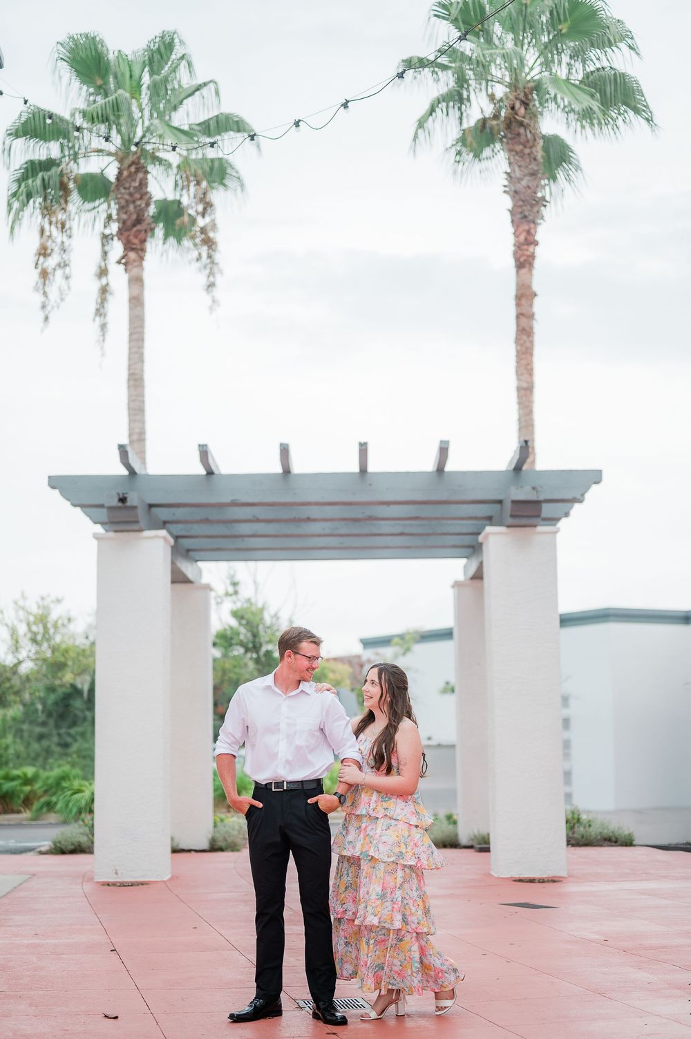 A romantic couple shares a tender moment together on a pink patio surrounded by tall palm trees in a tropical setting.