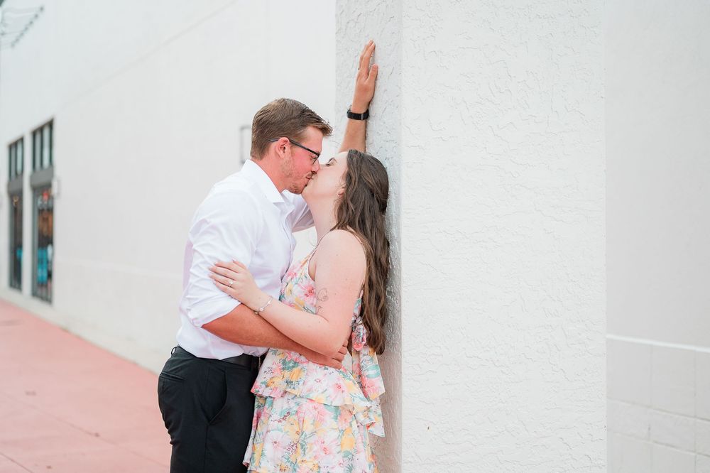 A couple shares a romantic moment against a white wall, with the woman wearing a floral dress and the man in business attire.