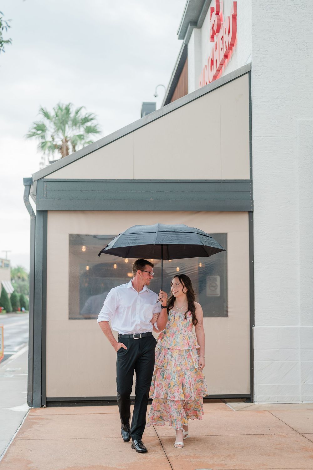 A couple shares romantic moments under a black umbrella in front of a modern building exterior.