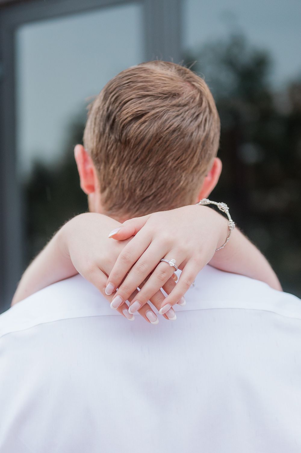 A close-up view of hands with an engagement ring clasped around a shoulder wearing a white shirt.