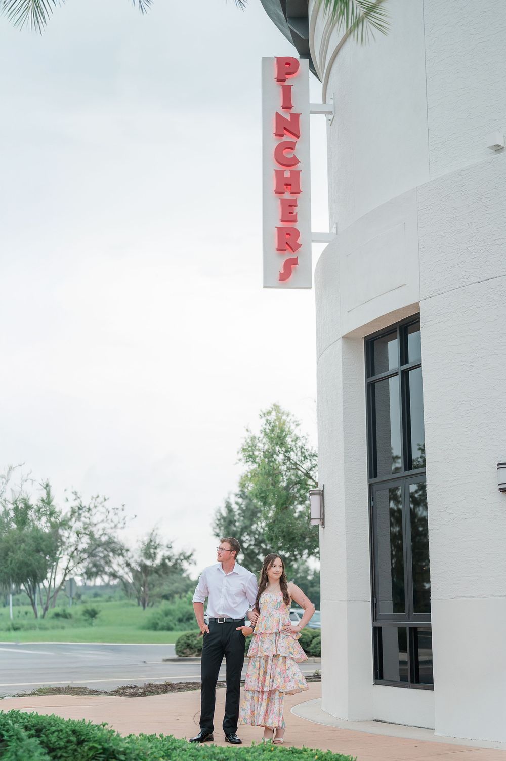 A couple stands outside a vintage-style building with a red 'Pichery' sign mounted vertically on the white exterior wall.
