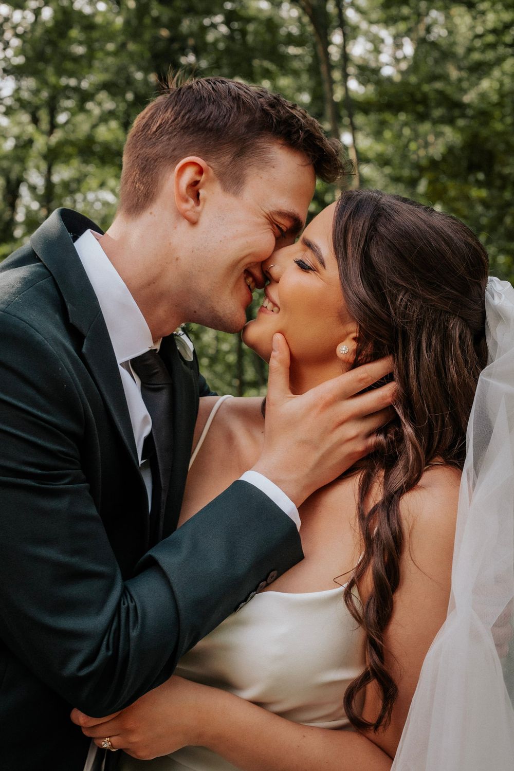 Bride and groom share romantic almost-kiss in lush green forest setting.