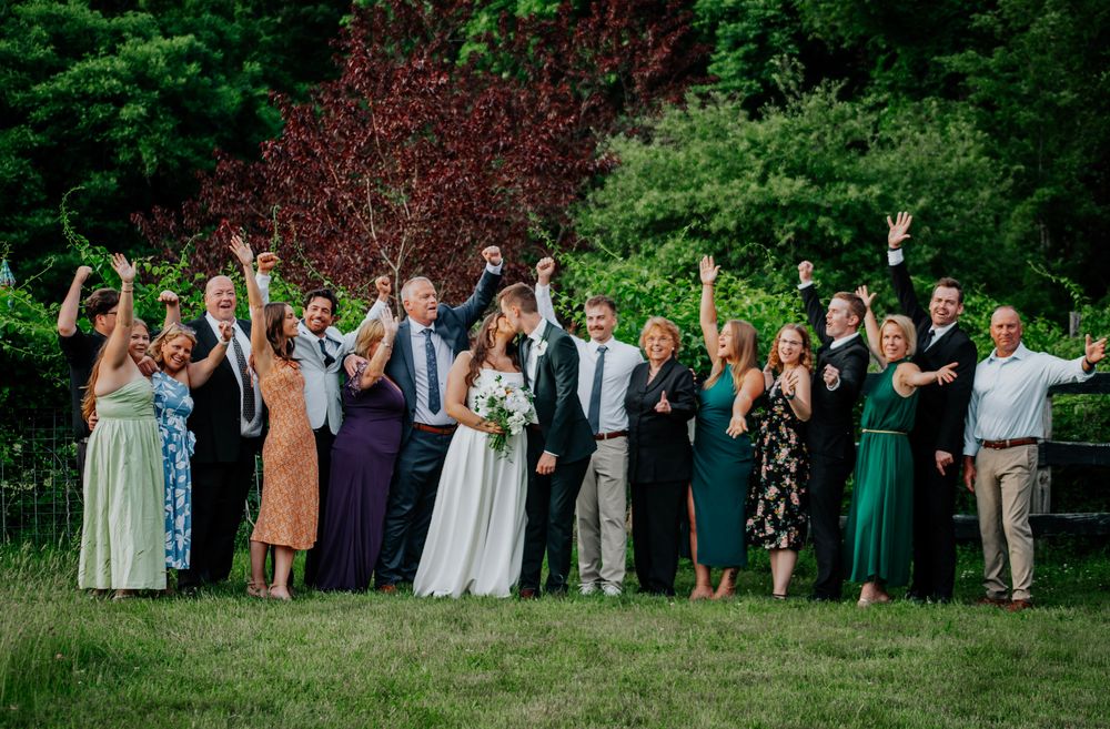 Wedding party celebrates outdoors with raised arms in lush green garden.