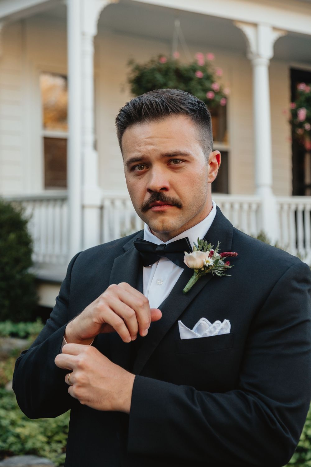 Dapper groom in black tuxedo with boutonniere adjusts his cufflinks outside a Victorian-style home.