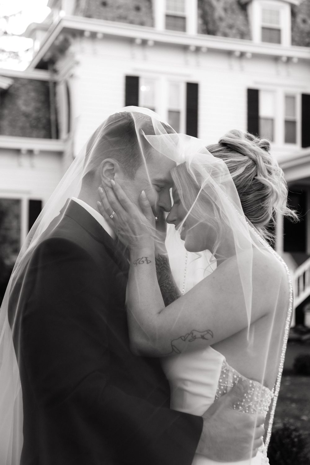 Bride and groom share an intimate moment under a wedding veil in black and white portrait.