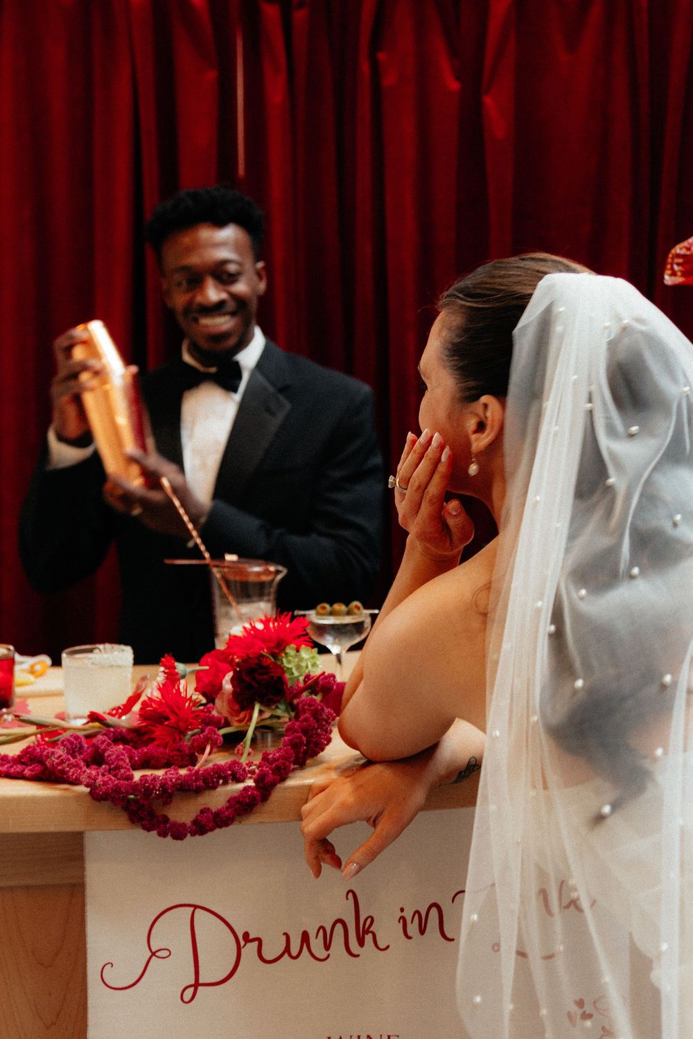 Bride laughing at bar while groom pours cocktail shaker at elegant red-themed wedding reception.
