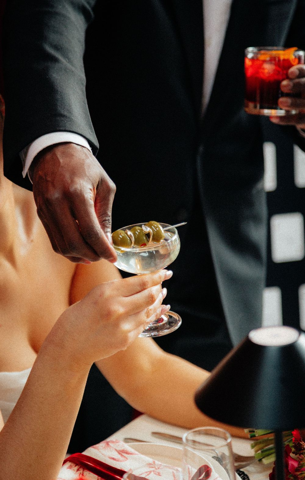 Groom handing bride a martini with olives at elegant wedding reception table.