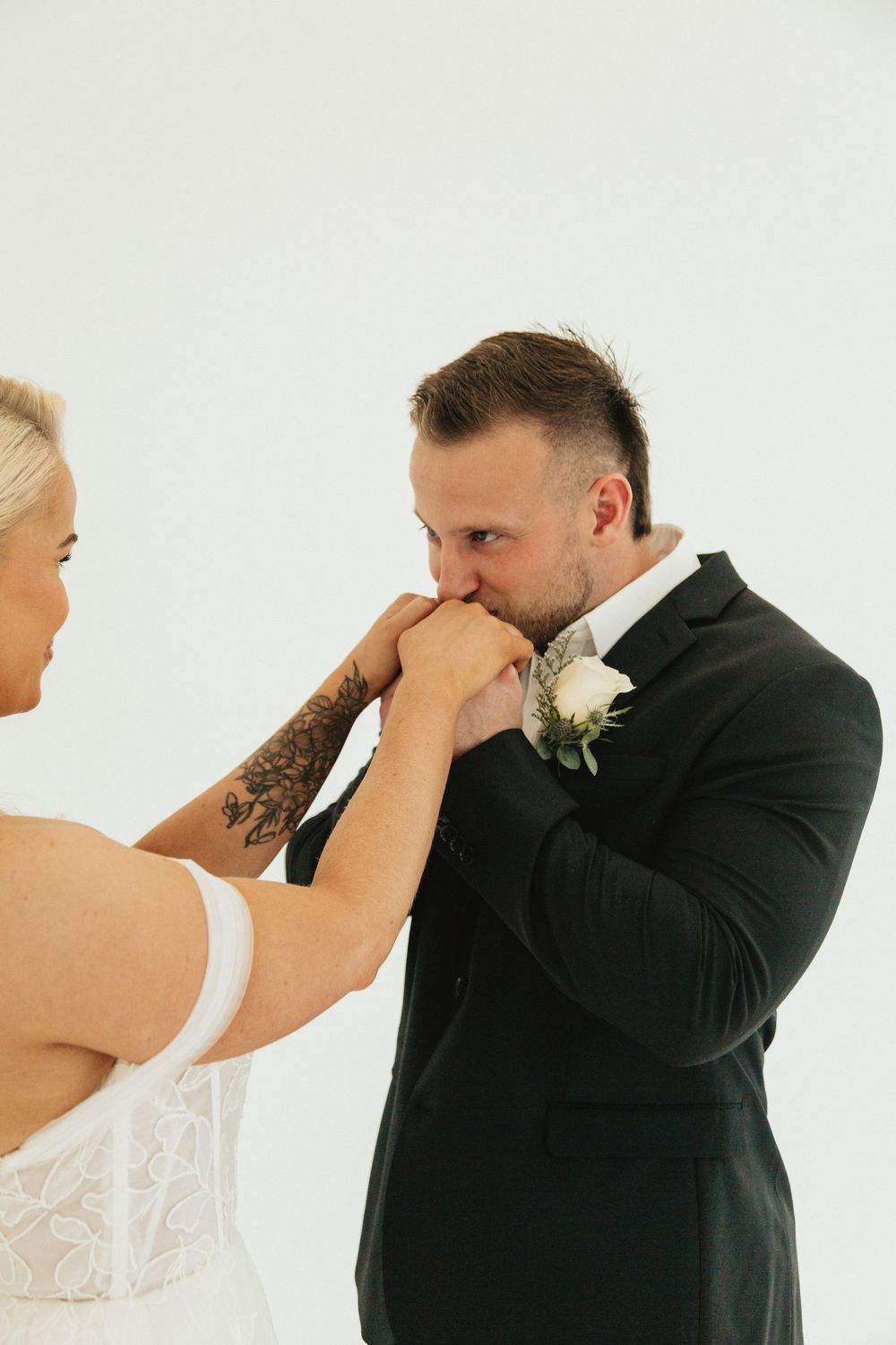 Groom in black suit tenderly kisses bride's hand during romantic wedding moment against white background.