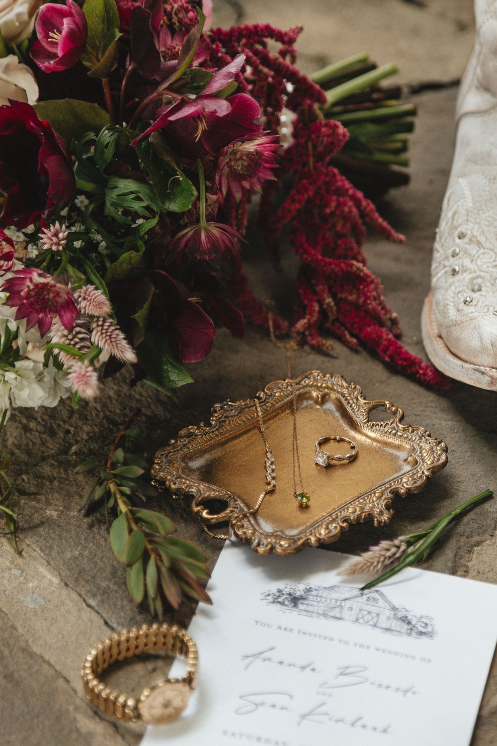 Wedding flat lay with gold jewelry tray, burgundy bouquet, invitation, and bridal accessories.