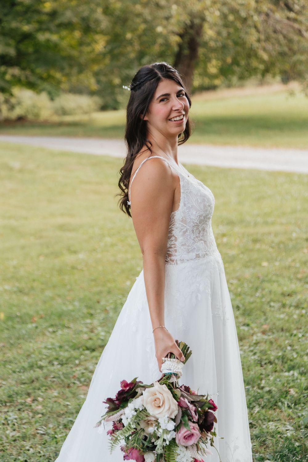 Smiling bride in lace wedding gown holding burgundy and blush bouquet outdoors.