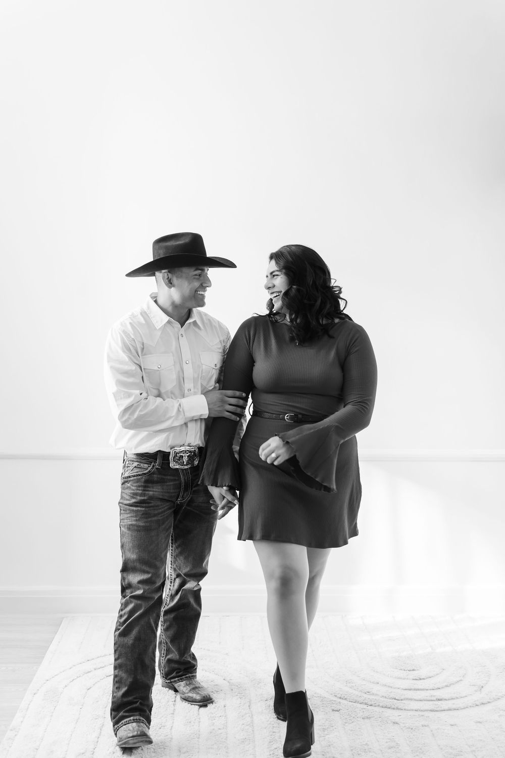 A couple in western attire walking together and smiling in a black and white portrait studio setting.