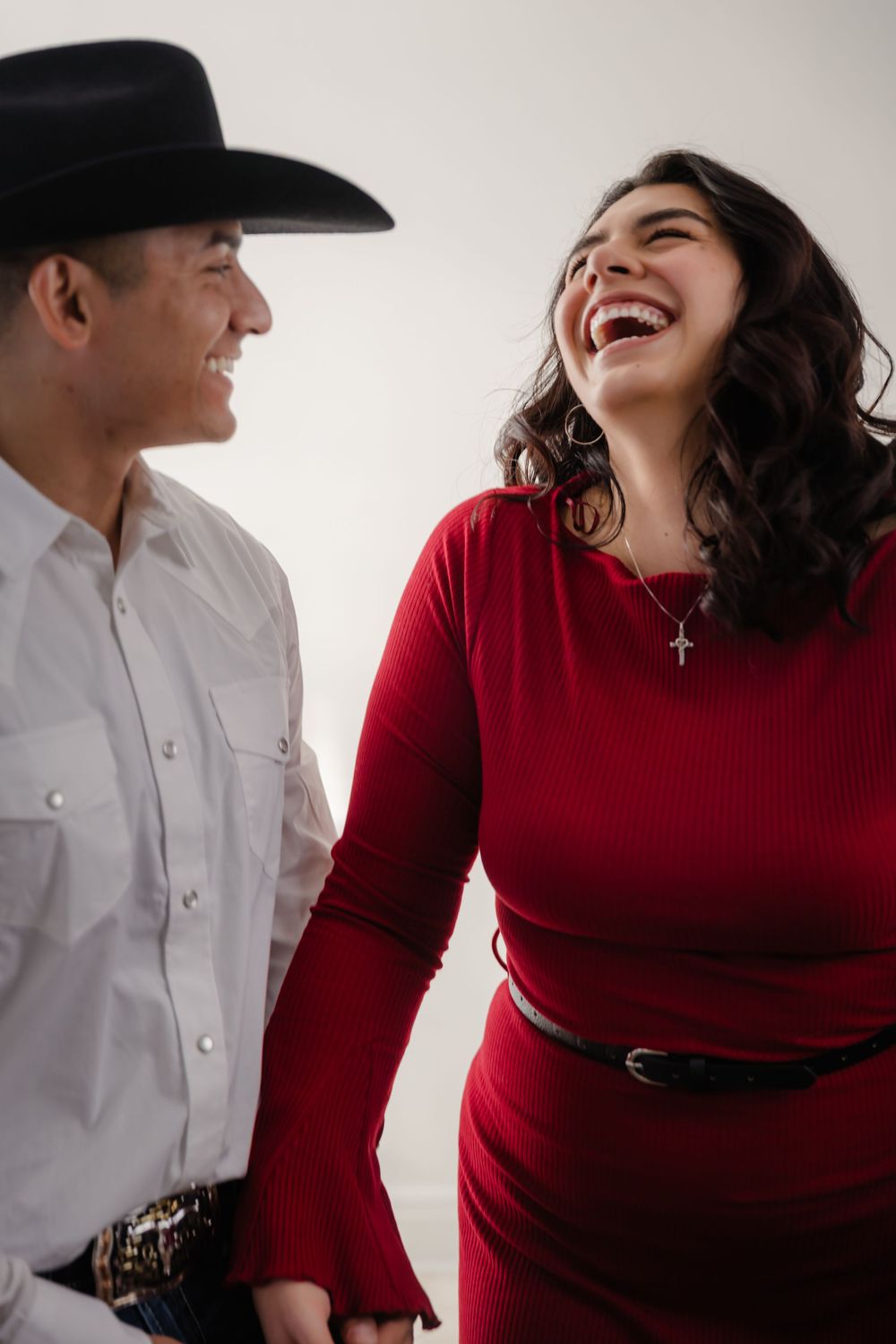 A couple in western attire, with a person in a white shirt and cowboy hat beside someone wearing a red sweater, laughing together.