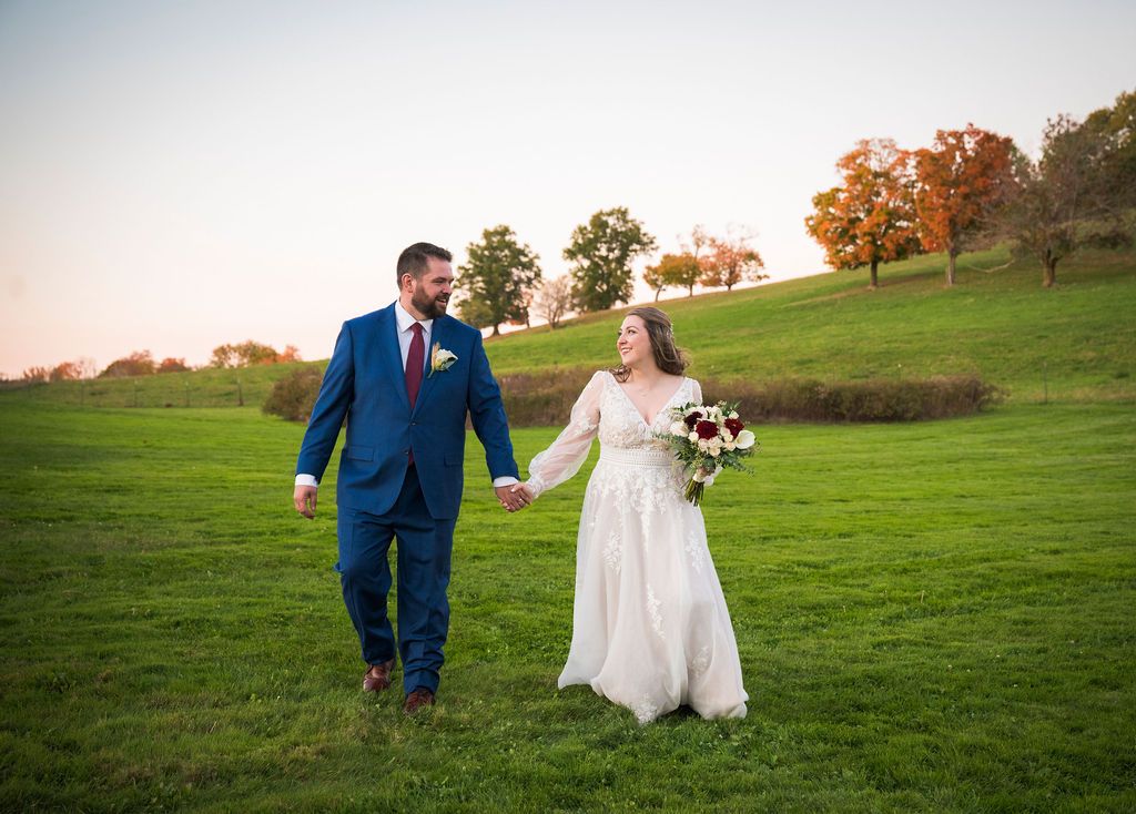 A couple in wedding attire walks hand in hand across a lush green hillside at sunset.