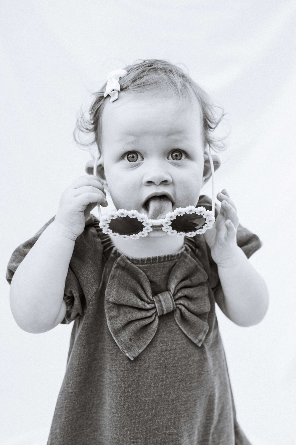 Cute little girl with a bow in her curly hair, playfully licks her flower-shaped sunglasses.