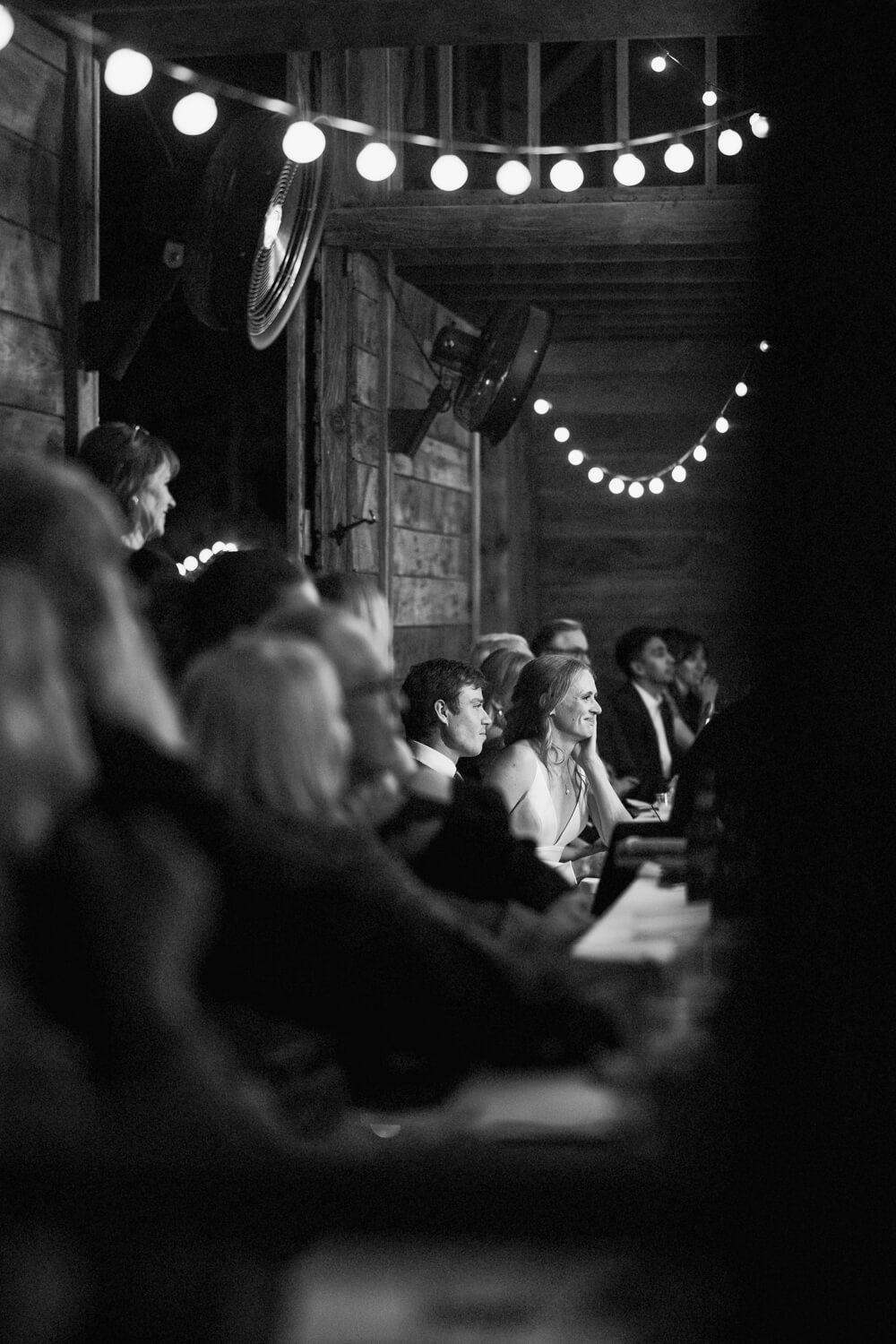 Bride and Groom surrounded by guests in a barn reception, listen to their best man give an emotional speech and toast.