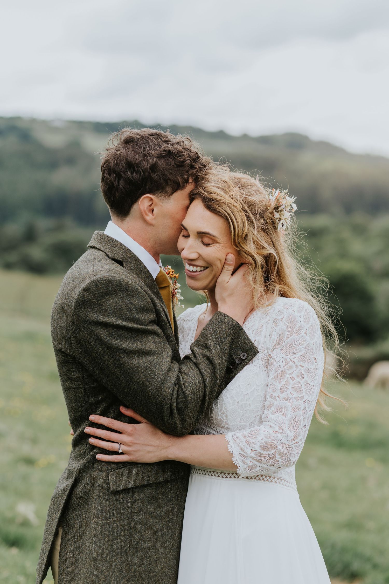 A romantic couple shares an intimate moment on a rocky beach landscape with a pink flower boutonniere accent.