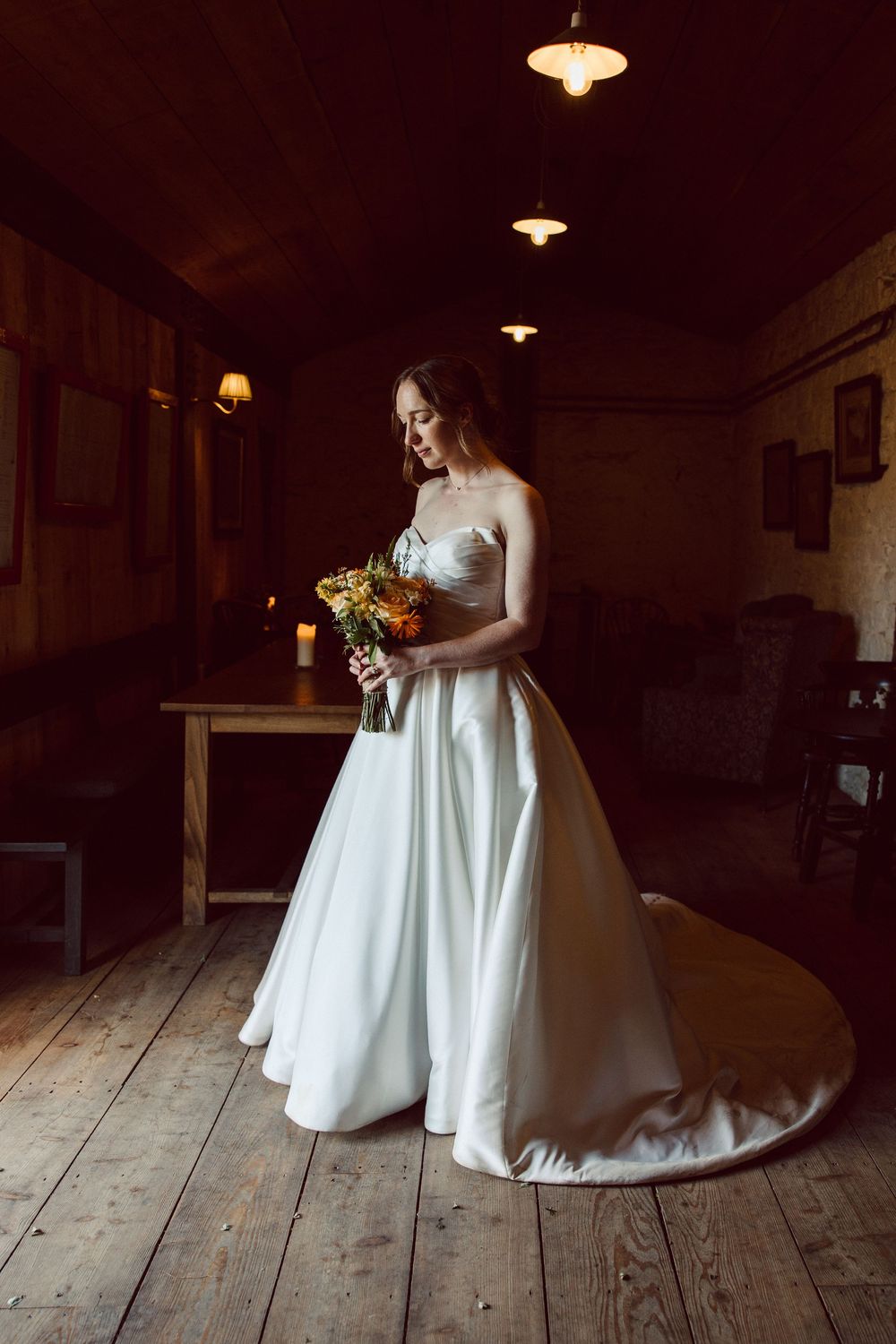 A bride in a classic white strapless wedding gown holds a sunflower bouquet while standing in a rustic wooden room.