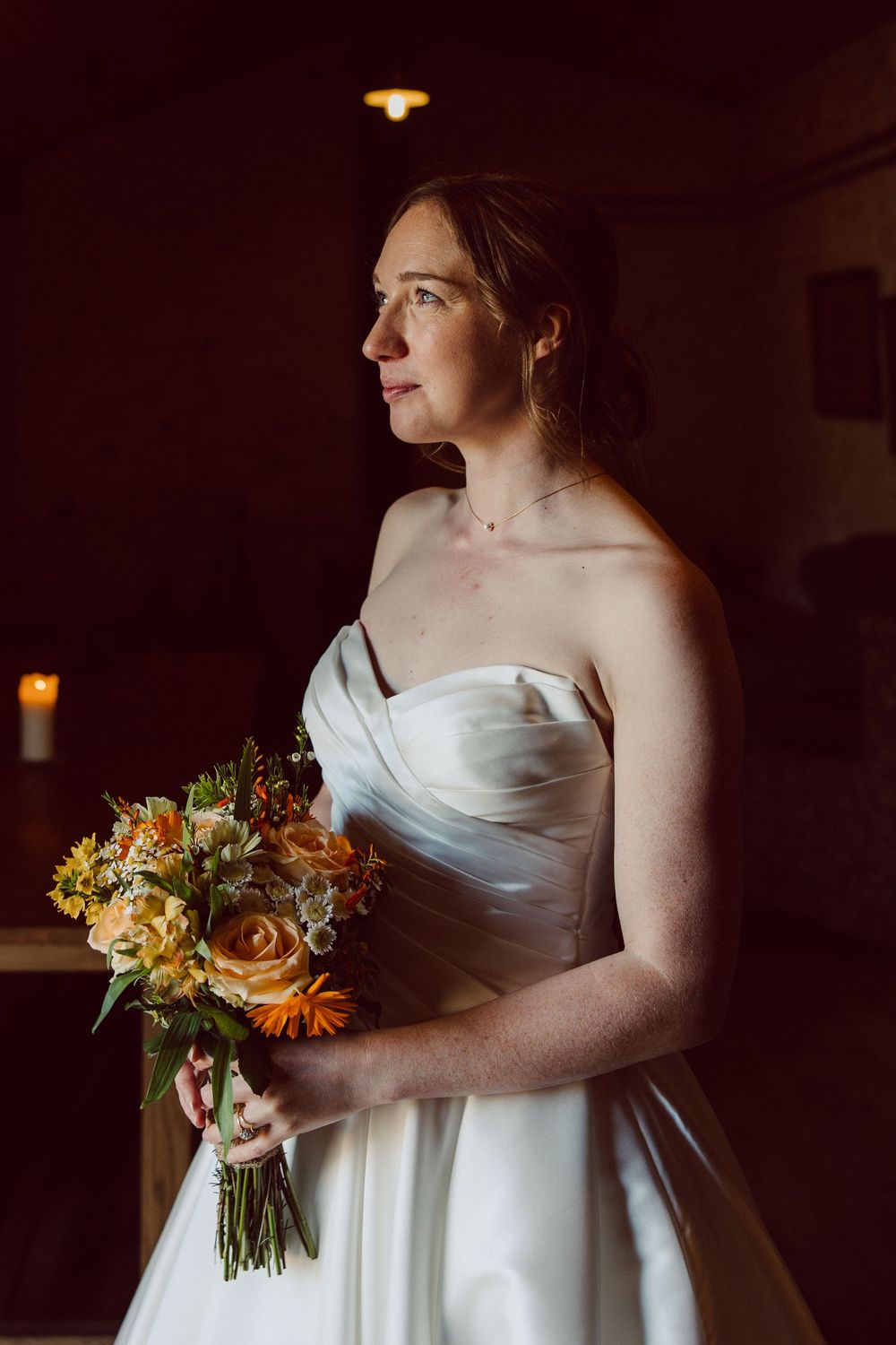 A bride in a white wedding dress holds a vibrant autumn bouquet of orange and yellow flowers in a dimly lit room.