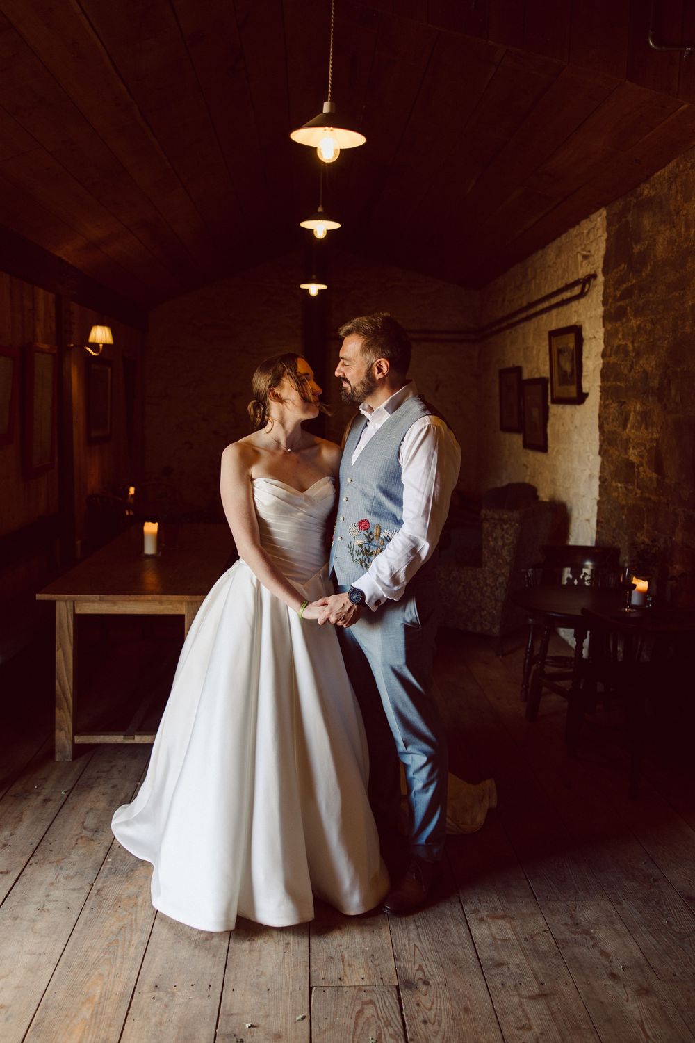 A couple in wedding attire dances together in a rustic barn venue with wooden floors and pendant lighting.