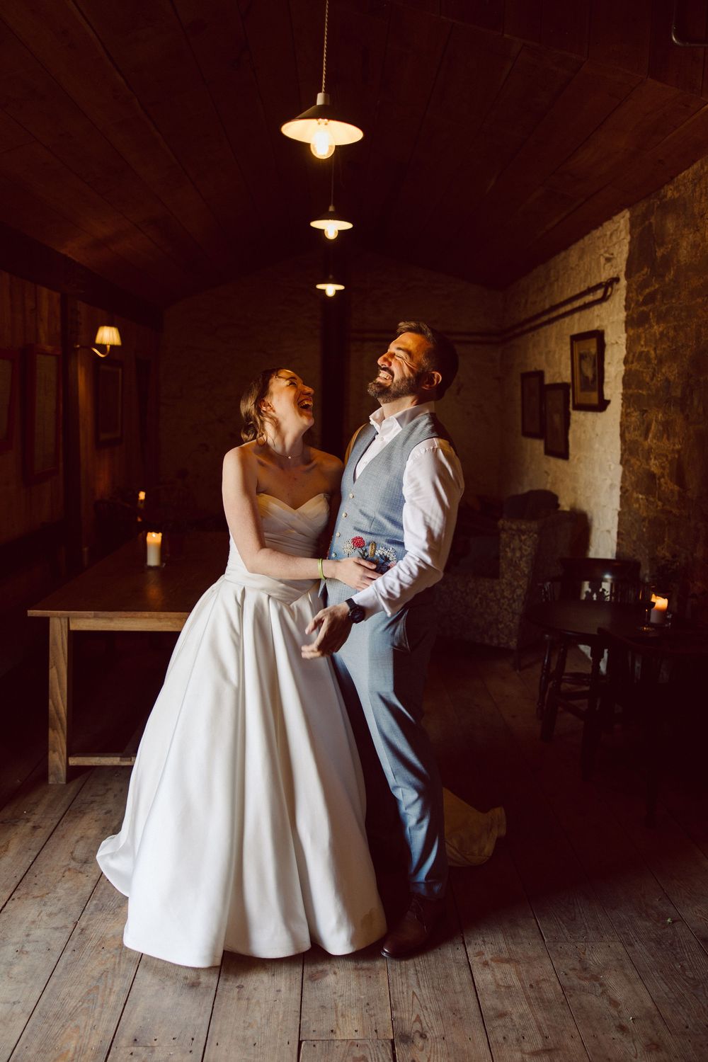 A couple in wedding attire dances together in a rustic barn setting with wooden floors and pendant lighting.