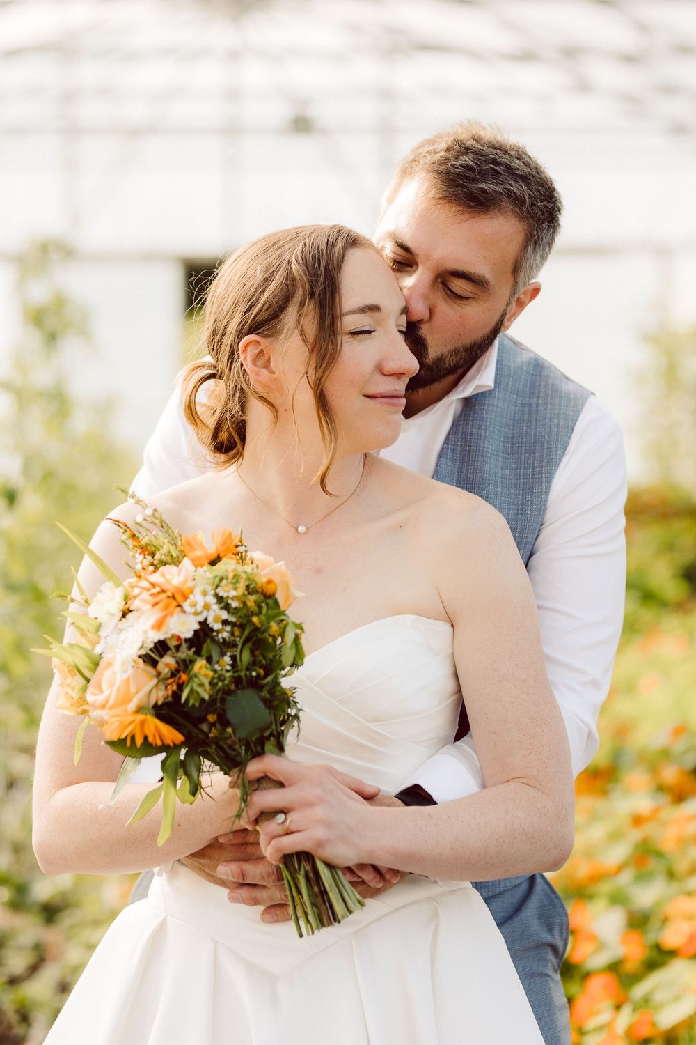 A romantic wedding couple embraces in a garden with orange flowers and a bridal bouquet.