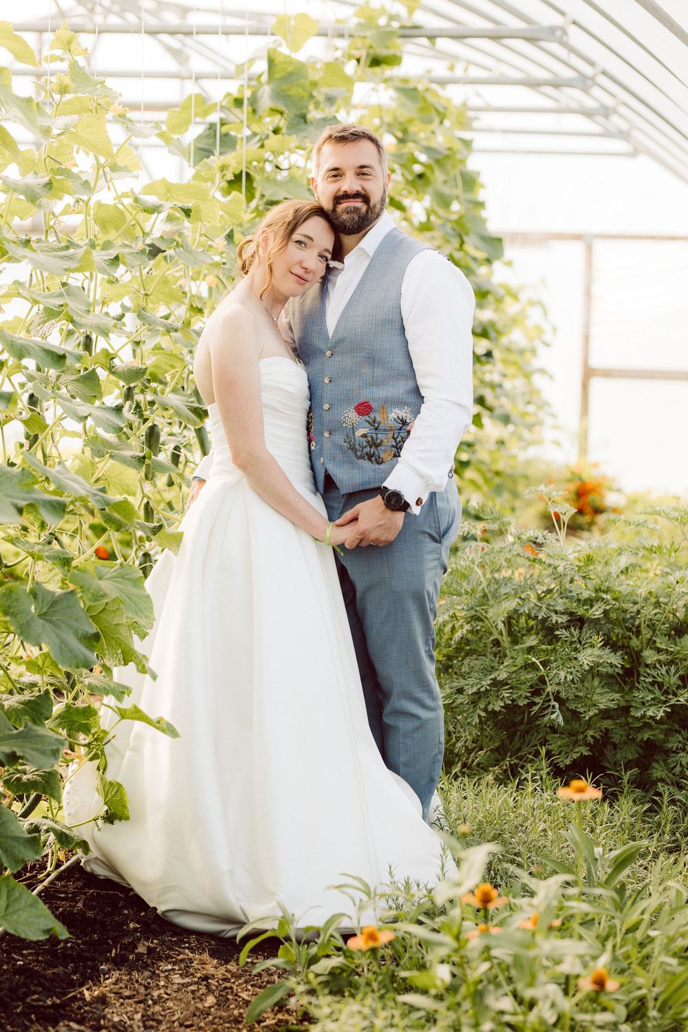 A romantic wedding couple embraces in a sunlit greenhouse surrounded by lush green plants and flowers.