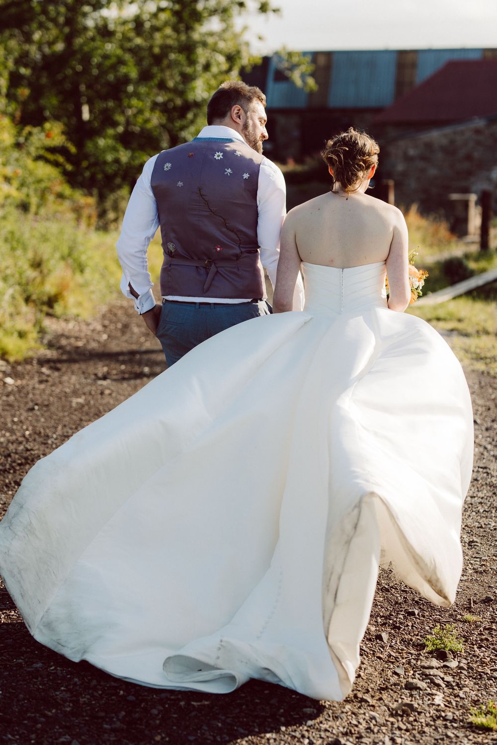 A couple in wedding attire walk together outdoors, with the bride's white gown flowing in a scenic natural setting.