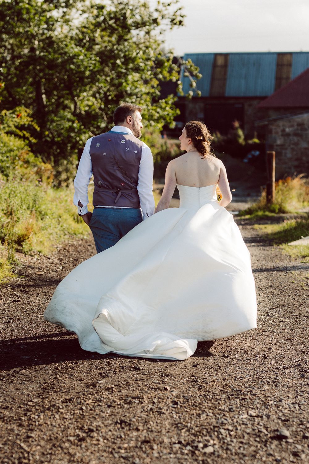 A wedding couple sits together on a gravel path during golden hour with their backs to the camera.