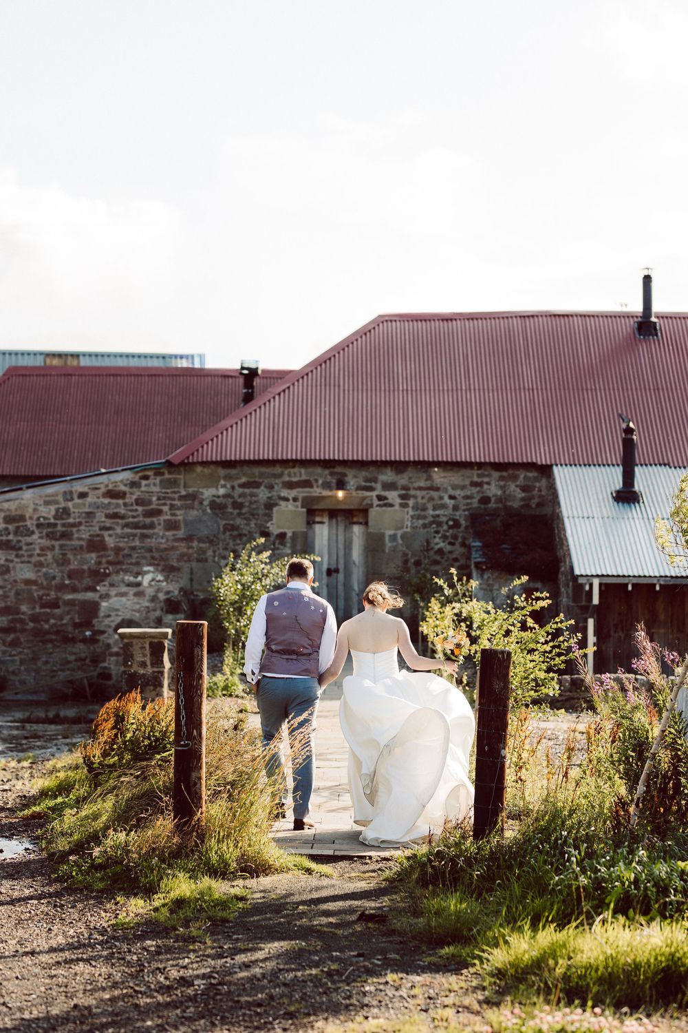 A wedding couple walks along a rustic stone building with sunlight casting long shadows.