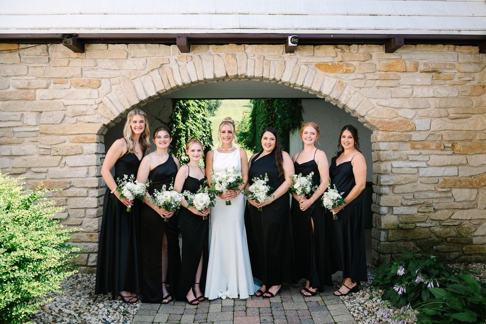 Wedding party wearing black and white formal attire poses together against stone wall backdrop.