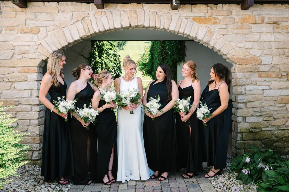 A wedding party in black and white dresses poses together against a stone wall backdrop in a garden setting.