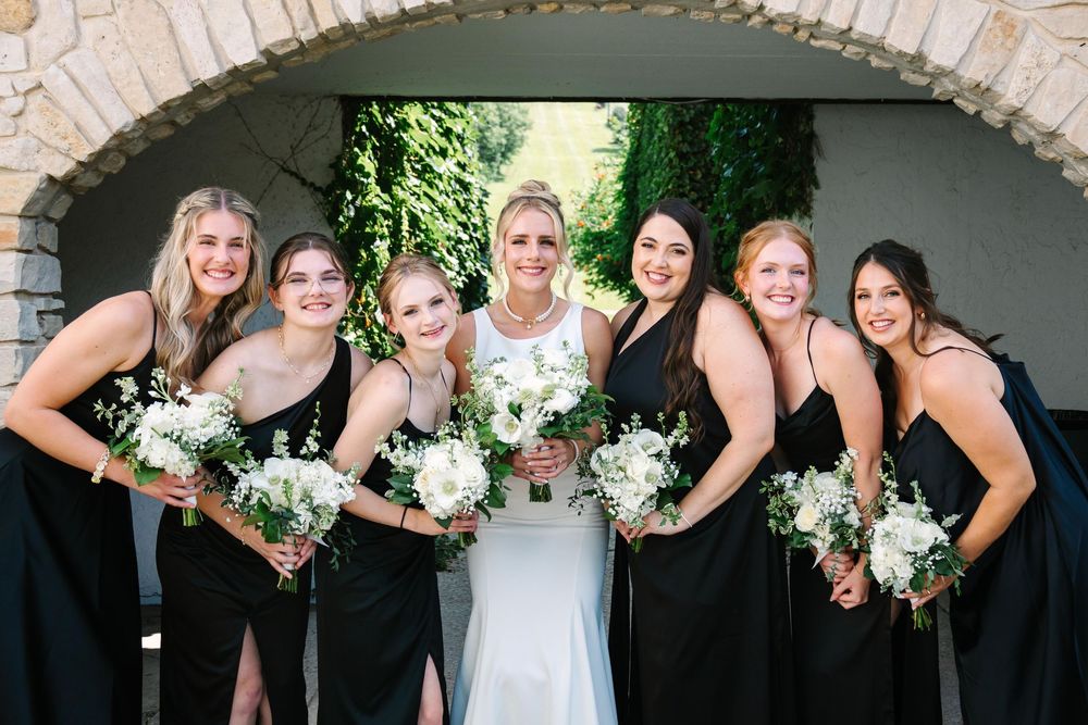 Wedding party in black dresses and white gown pose under stone archway holding elegant white floral bouquets.