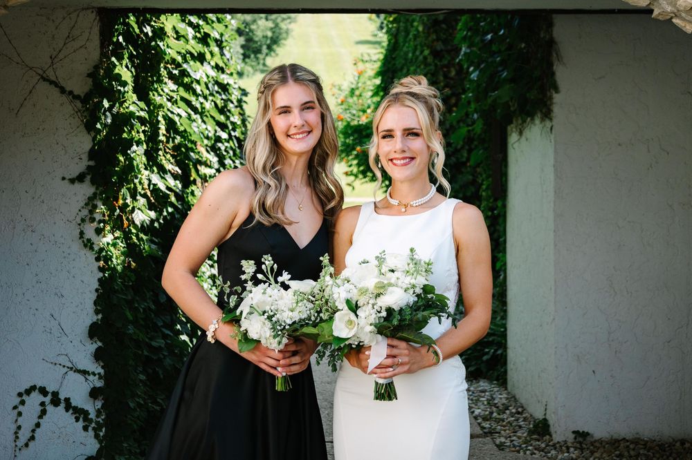 Two people in white dresses hold bouquets while standing together against a green vine-covered wall.