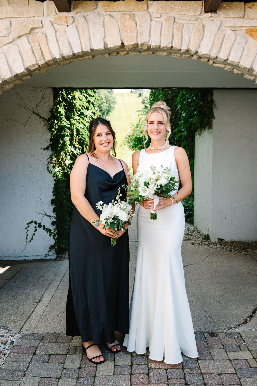 Two people in black and white dresses stand together under a stone archway in an outdoor courtyard setting.