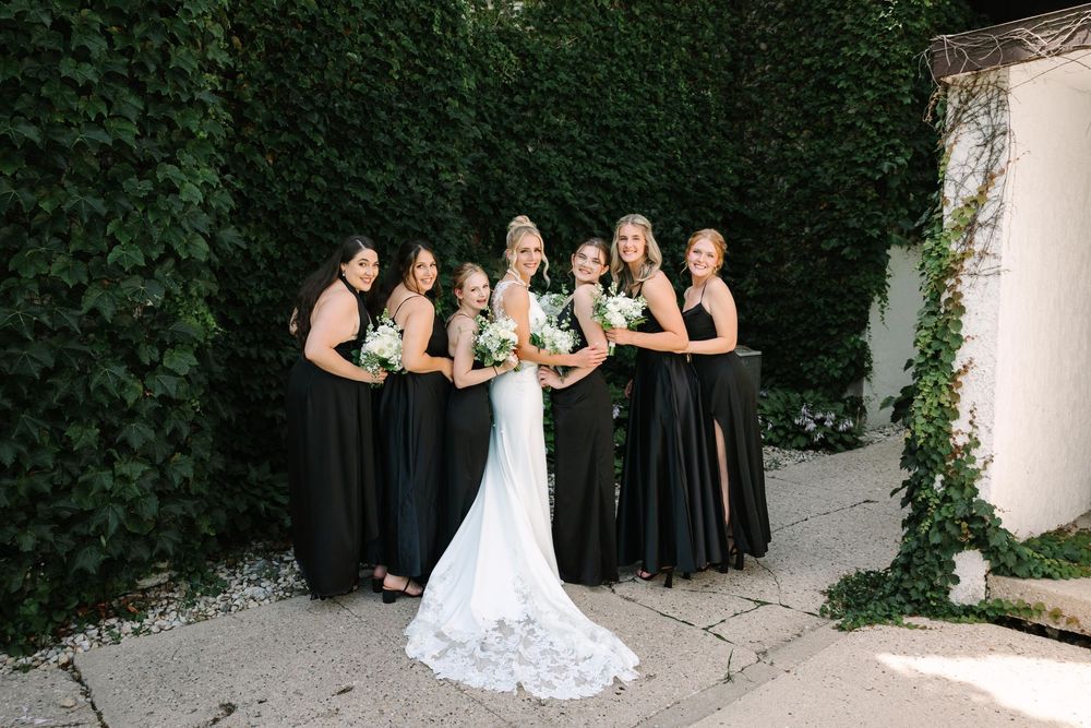 Wedding party poses together in front of lush green hedge wall wearing black dresses and white gown.