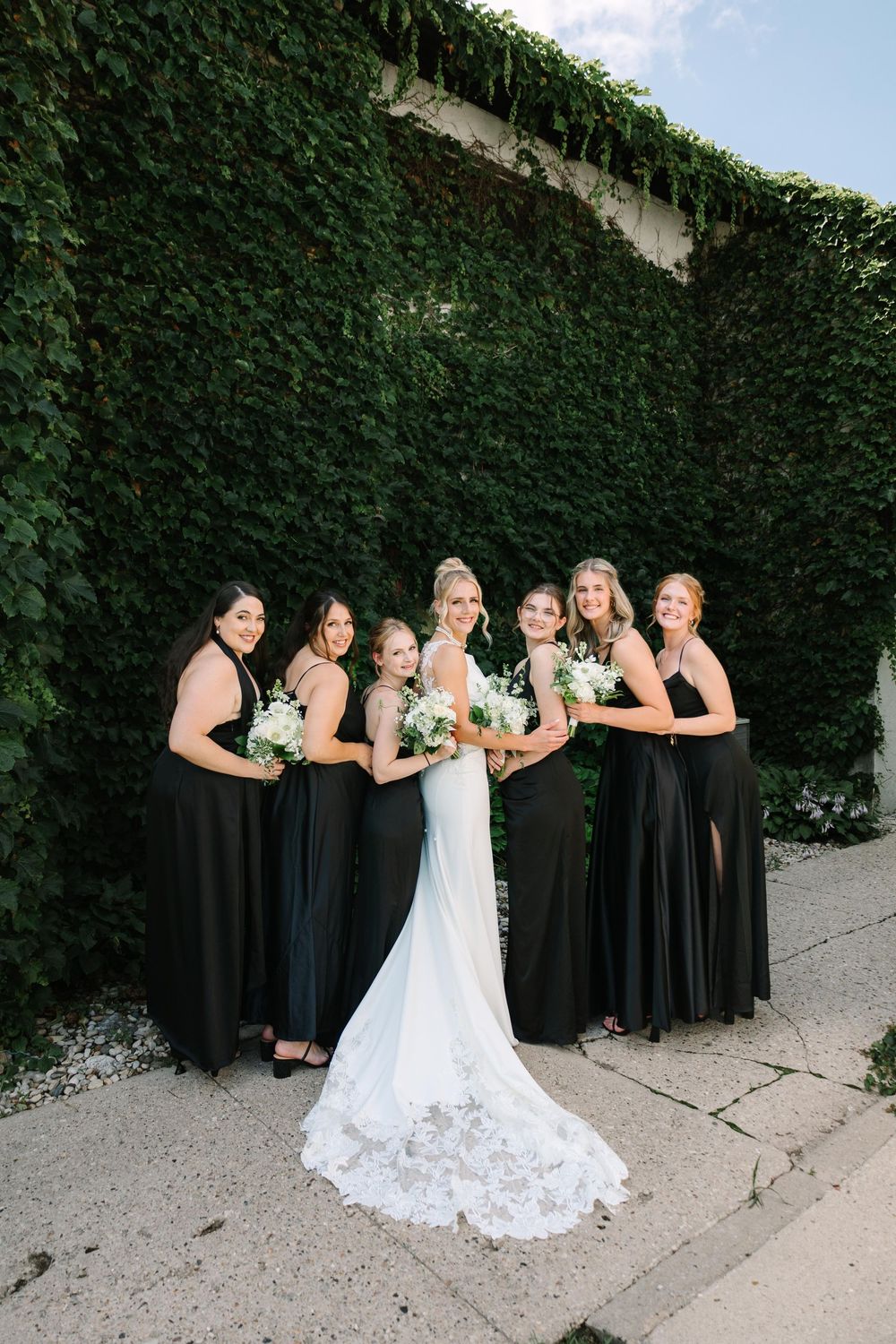 A wedding party poses together against a lush green ivy wall backdrop wearing black bridesmaid dresses and white bridal gown.