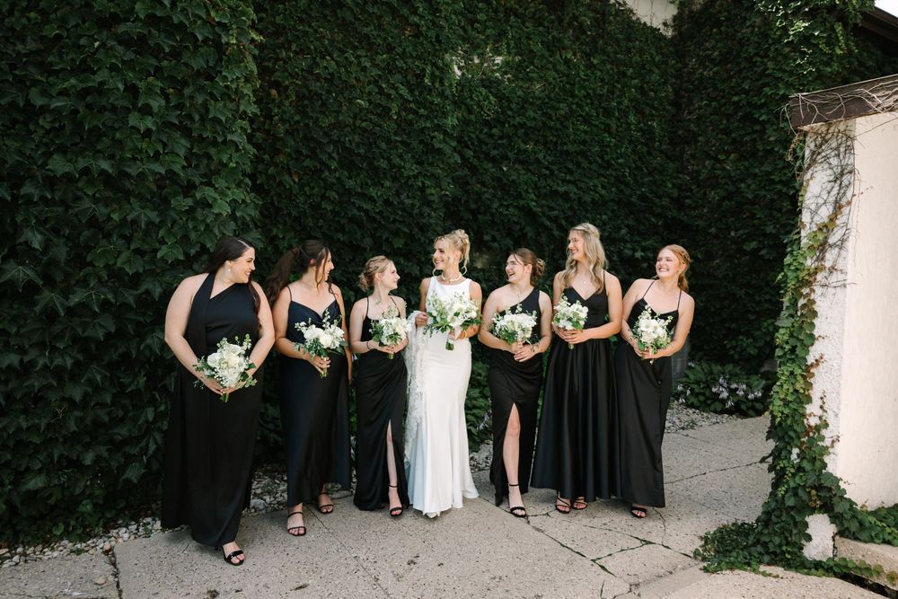 Bridal party in elegant black dresses poses against an ivy-covered wall during an outdoor wedding ceremony.