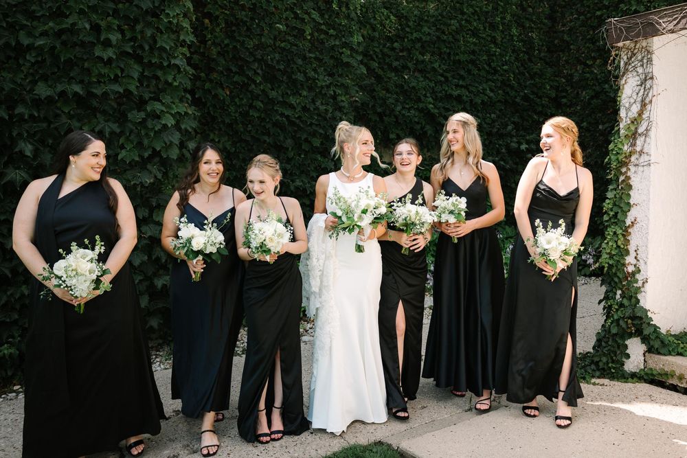 A bridal party wearing elegant black dresses and a white wedding gown pose together holding white floral bouquets.