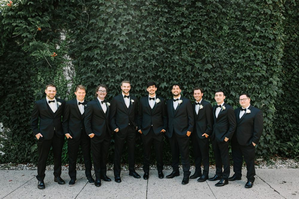 A group of groomsmen in black tuxedos stand in a line against an ivy-covered wall outdoors.