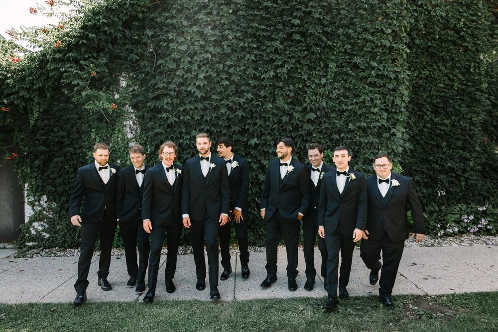 A group wearing black tuxedos and bow ties poses together outdoors against a green hedge backdrop.