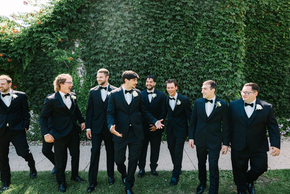 A line of groomsmen in black tuxedos and bow ties stand together against a green hedge backdrop at a formal event.