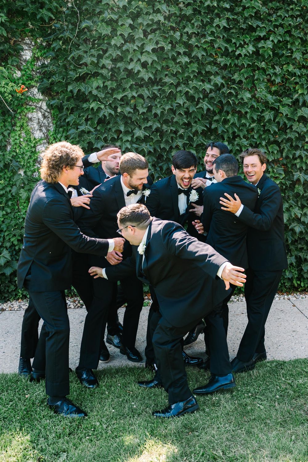 A group of groomsmen in black suits playfully fall over like dominos on grass in a sequence of candid wedding photos.