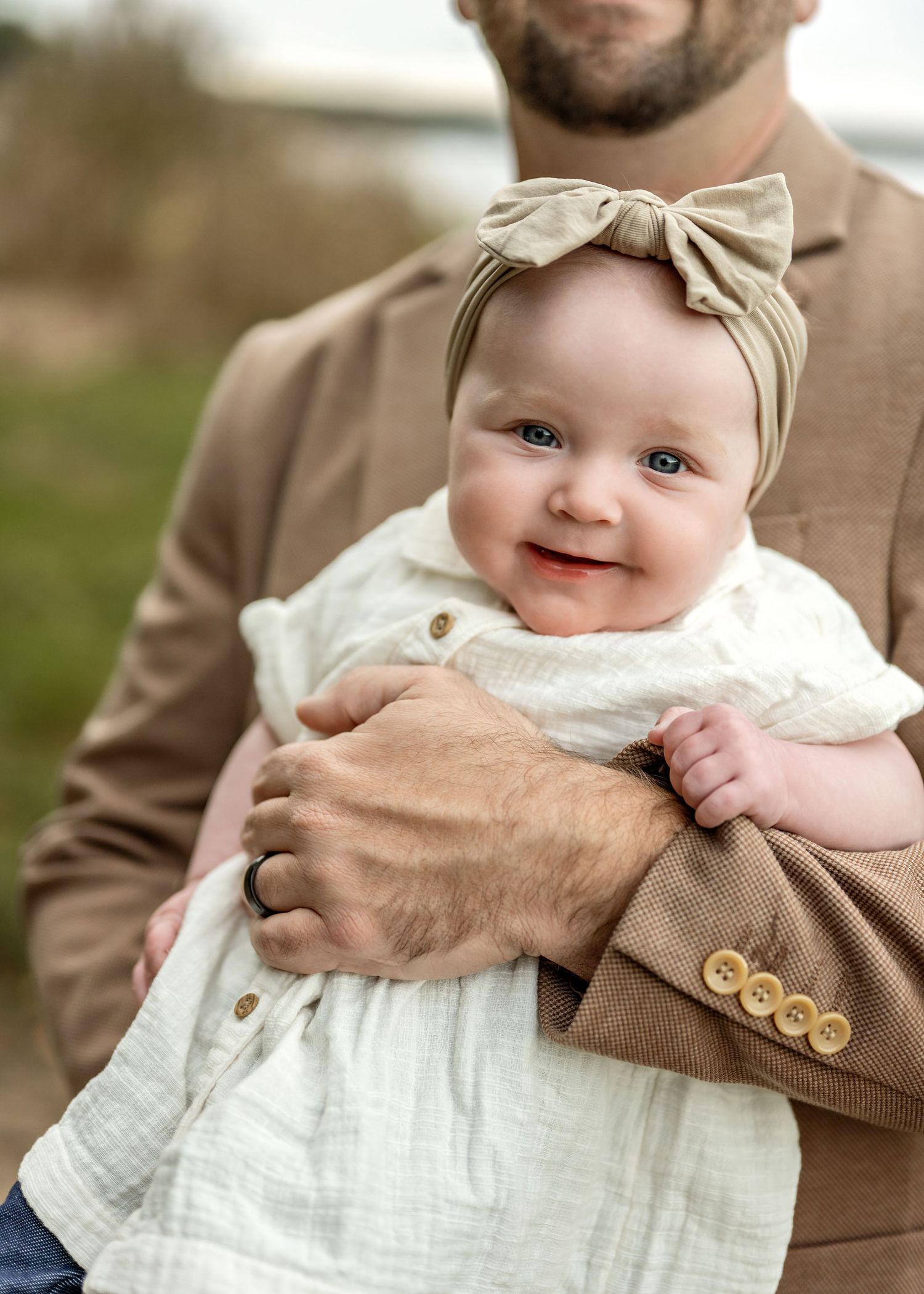 A series of close-up photos showing a baby in a white dress and tan bow being held in brown-clad arms.