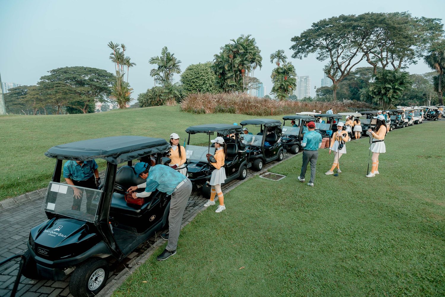 Golf carts lined up on a grassy course with players gathering for a tournament or event.